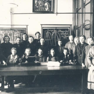 105-Toen er in 1923 een nieuwe meisjesschool gebouwd werd, gebruikte men de oude school overdag als kleuterschool en ‘s avonds als naaischool. Op deze foto uit 1926 zien we de leidster met haar leerlingen van deze naaischool, die maar een paar jaar heeft bestaan.